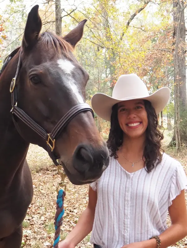 Smiling woman in a cowboy hat standing beside her brown horse in autumn woods