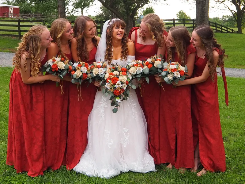 Laughing bride surrounded by bridesmaids in red dresses holding bouquets on a farm lawn